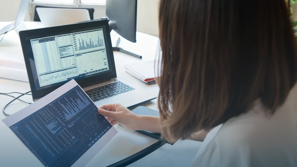 Analyst comparing a printed dashboard with charts on a laptop
