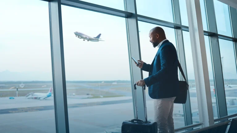 Business traveler with a suitcase checking his phone at an airport window as a plane takes off