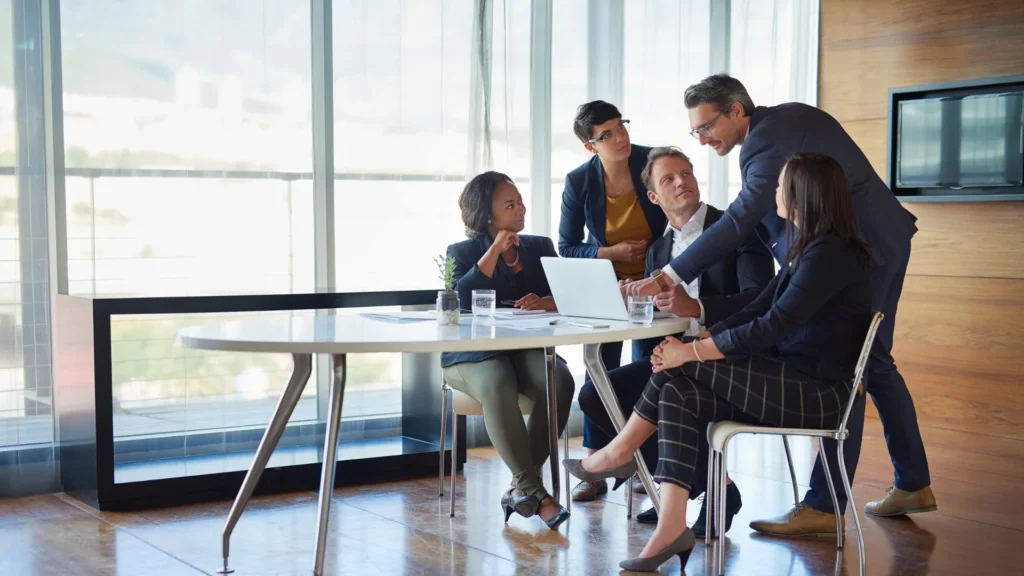 Business team gathered around a laptop in a glass-walled meeting room